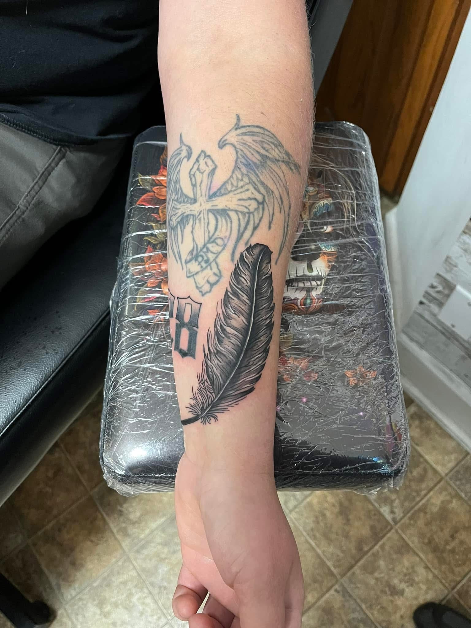 Winged cross with feather and number 8 forearm tattoo